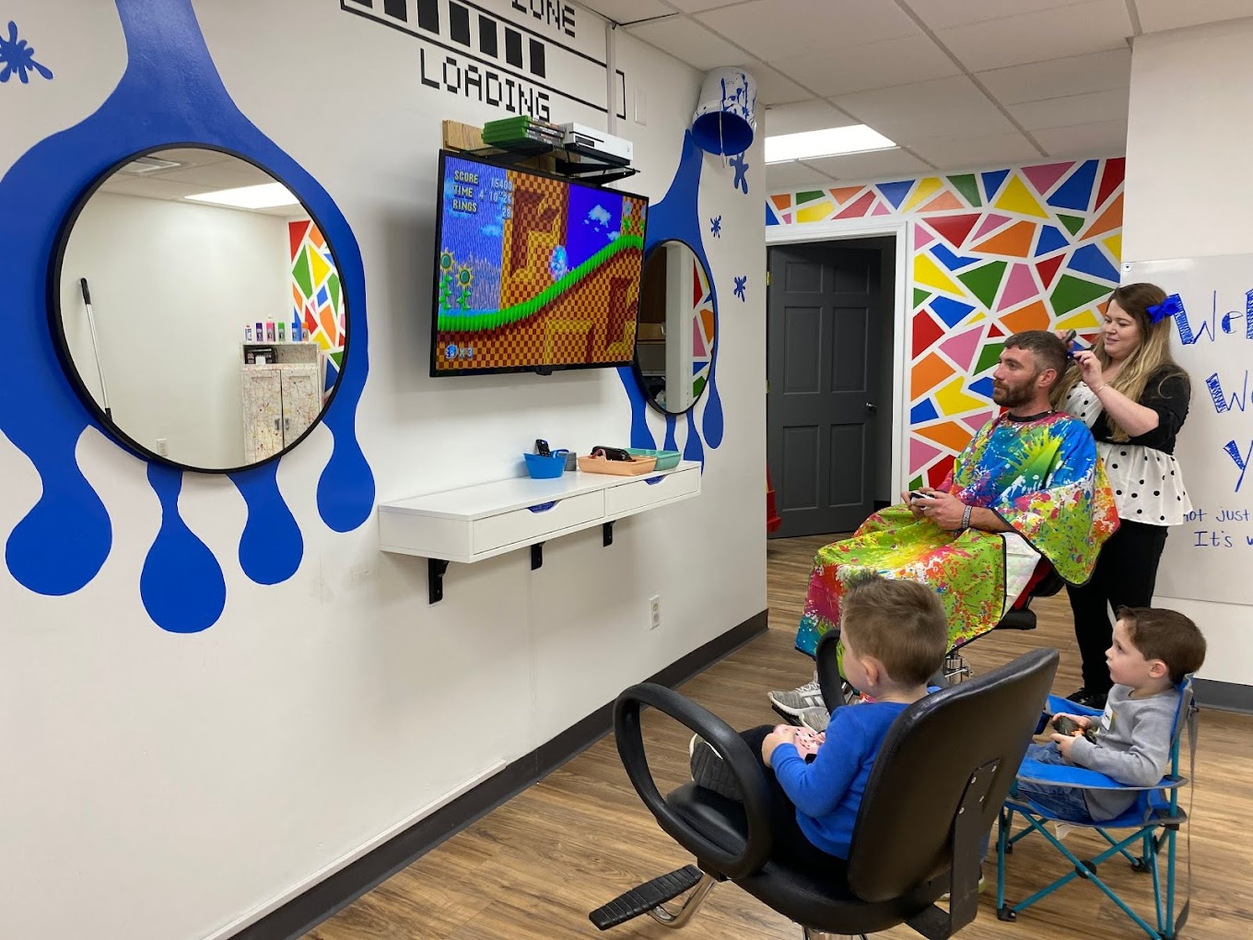 The salon interior: a wall painted with enormous blue paint drips with round mirrors set inside them, a screen and two game stations, and a man in a paint-splatter cape having his hair cut beside a wall of coloured triangles.