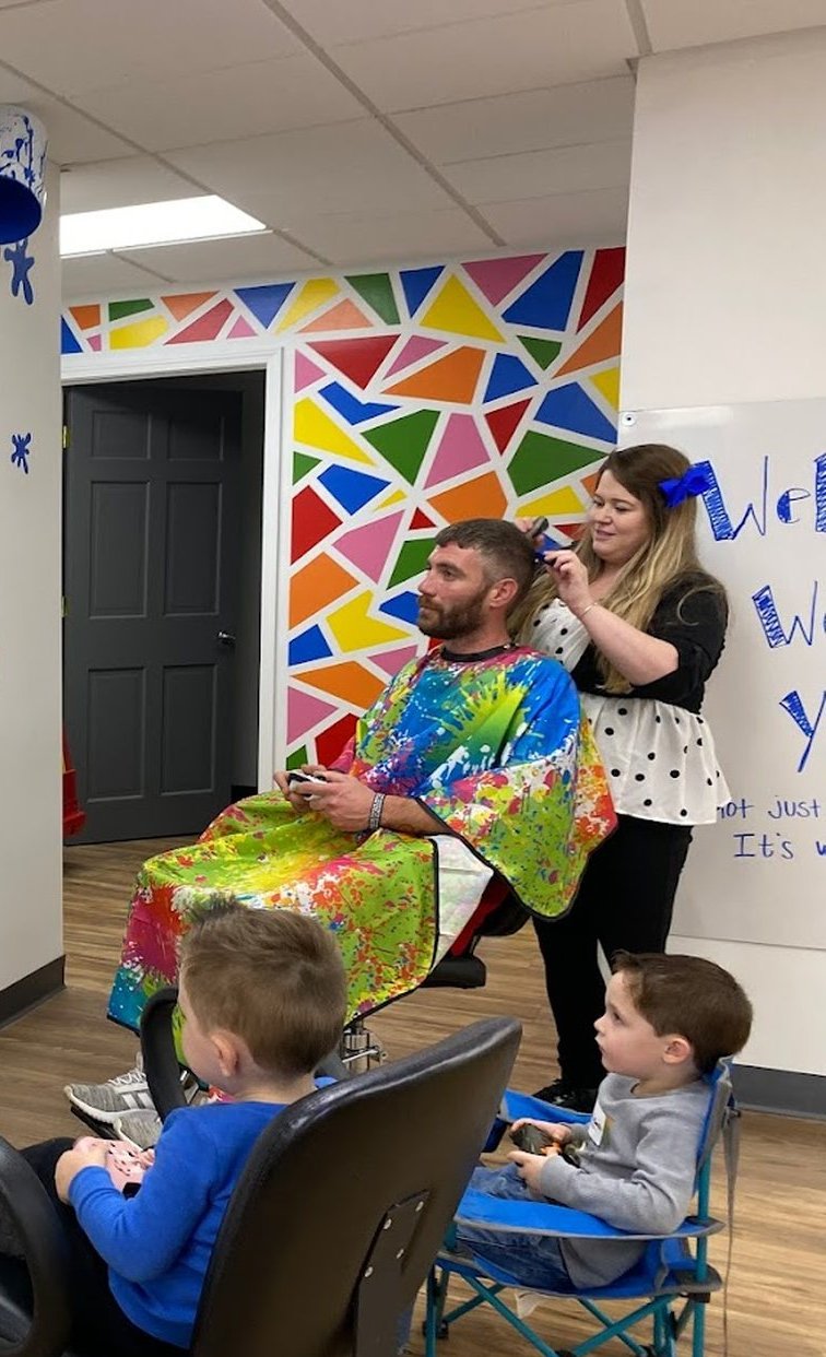 A wall covered in bright painted triangles beside a man in a rainbow splatter cape having his hair cut.