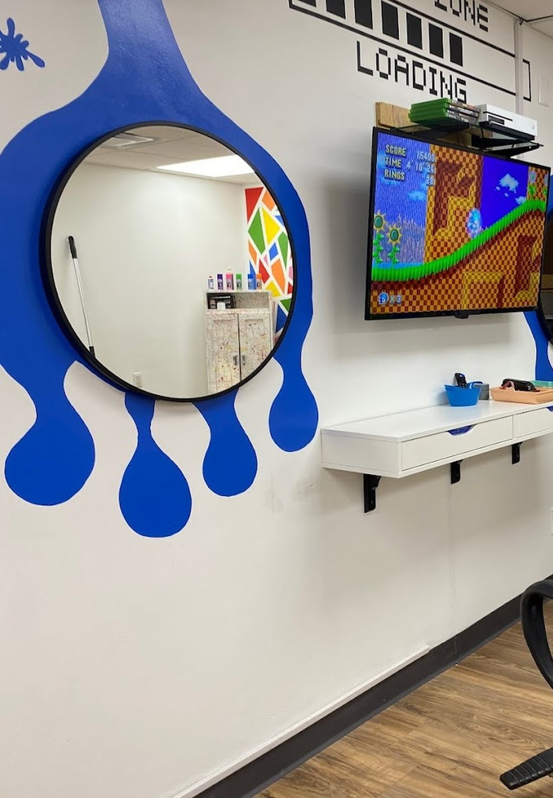A round mirror mounted inside a huge blue painted drip, with a screen and game console on a shelf beside it.