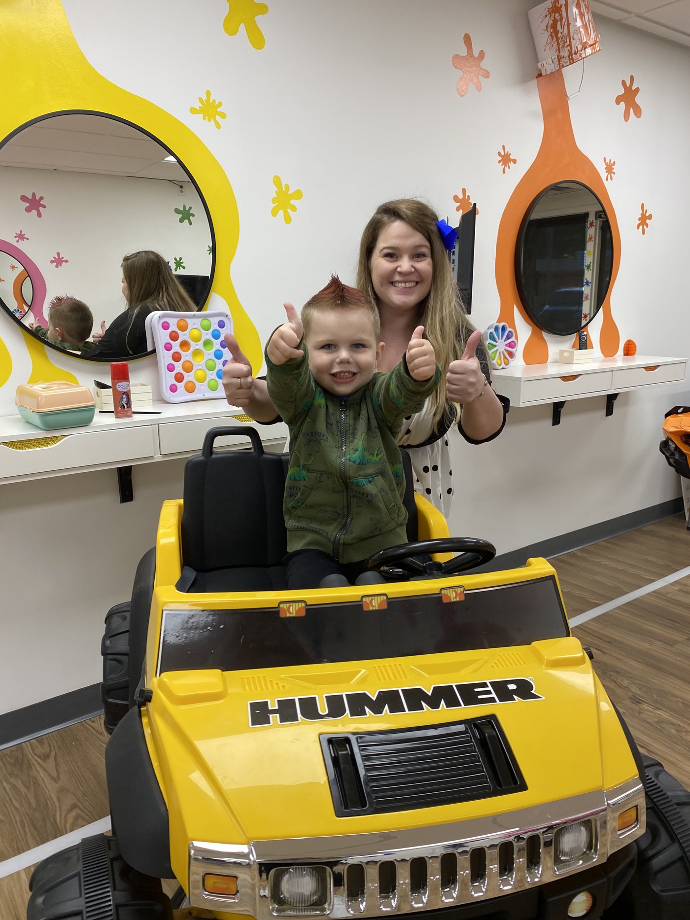 A yellow ride-on truck at a styling station, round mirrors set into orange and yellow splats on the wall behind.