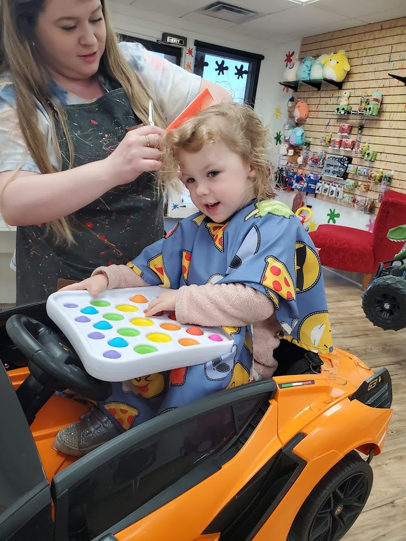 A stylist in a paint-covered apron trimming a toddler's hair in an orange ride-on car, the toy wall behind them.