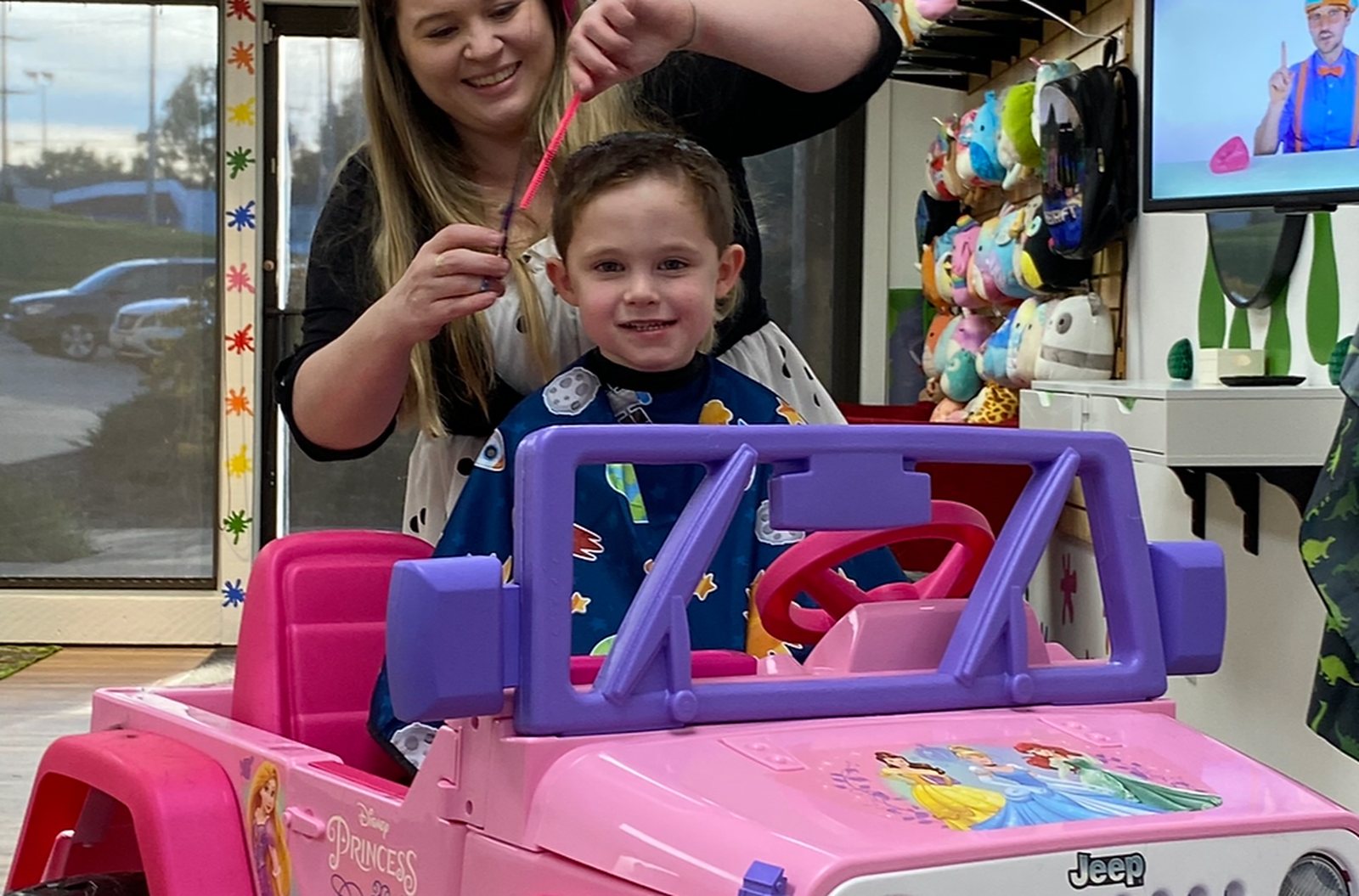 A child in a pink ride-on car by the front window while a stylist combs his hair, splat decals stuck across the storefront glass.