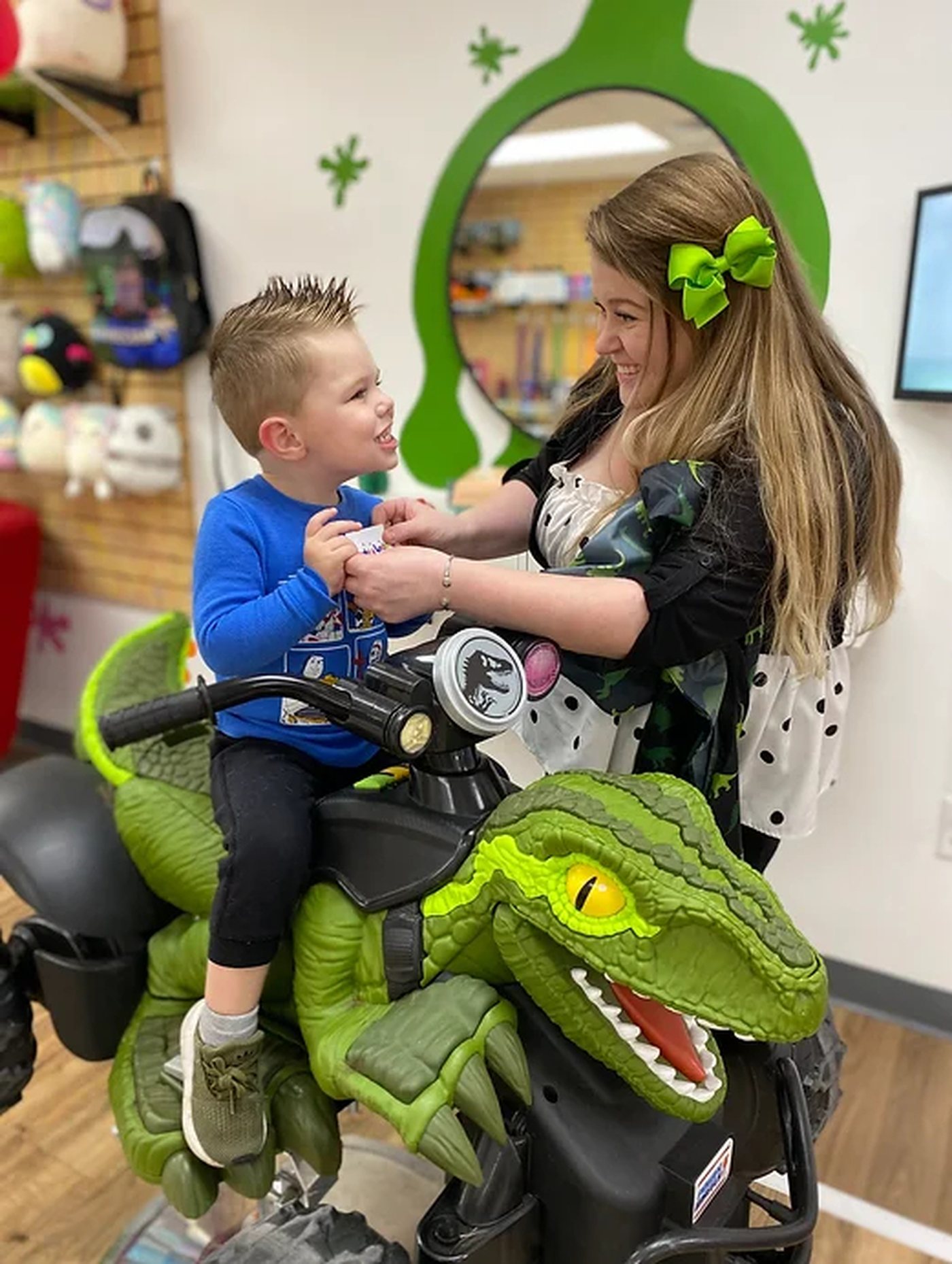 A boy on a large green ride-on dinosaur laughing with his stylist, in front of a green painted splat.